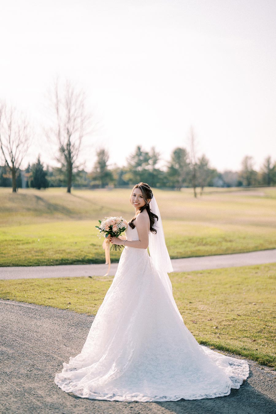 A bride in a white wedding dress holding a bouquet stands outdoors on a sunny day, with a grassy landscape and trees in the background.