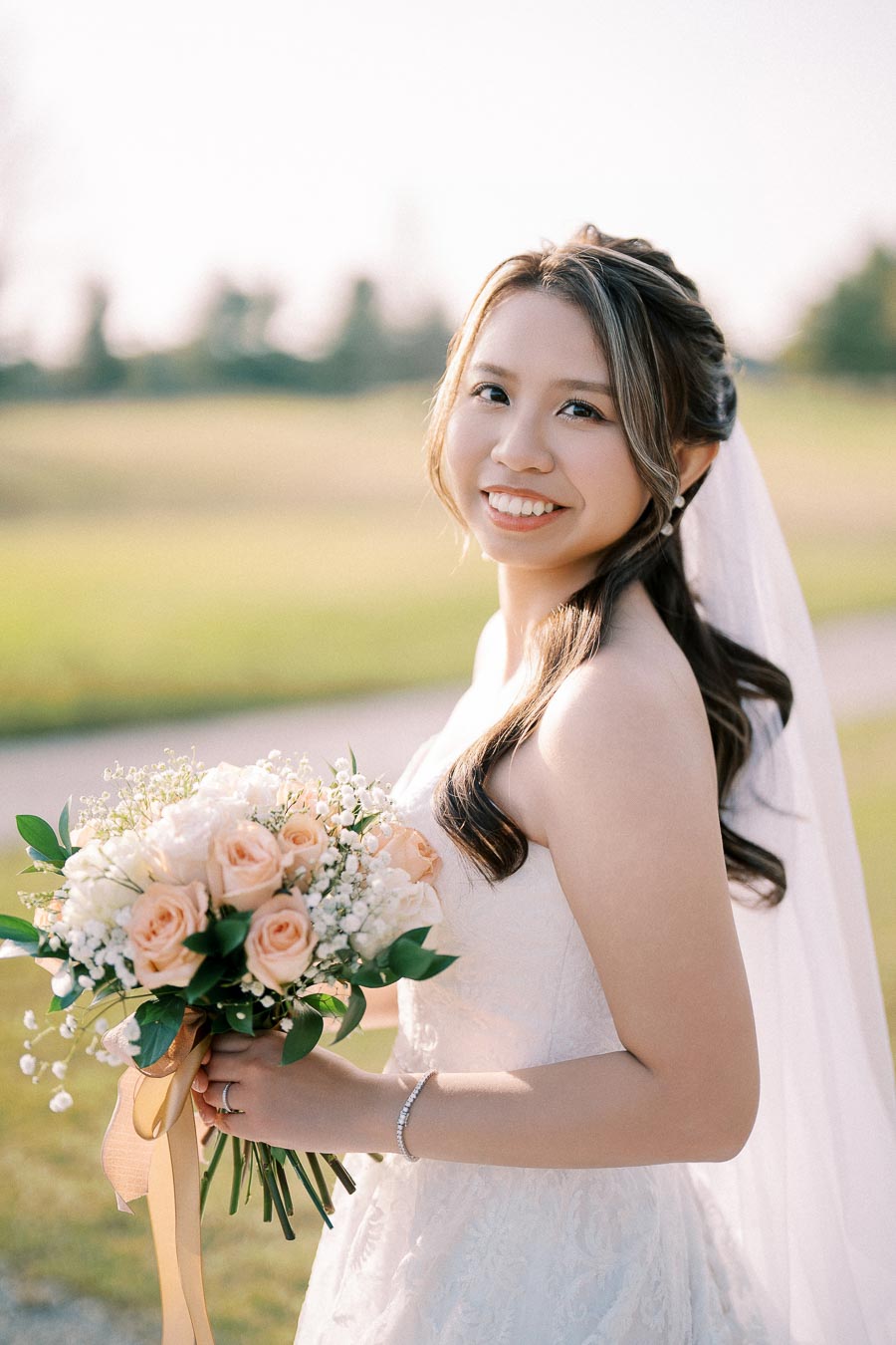 A bride in a white wedding dress holding a bouquet of peach roses and white flowers, smiling outdoors on a sunny day.