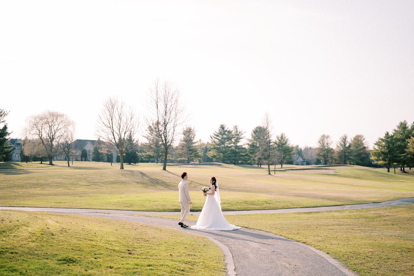 Wedding couple walking on a scenic golf course path, surrounded by green grass and trees, with a clear sky above.