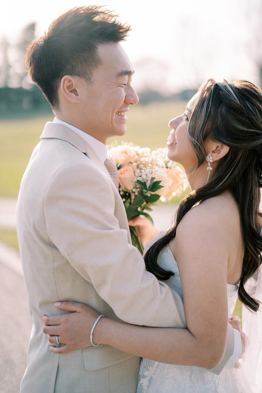 A happy couple embracing at their outdoor wedding, with the bride holding a bouquet of roses and both smiling joyfully against a blurred green background.