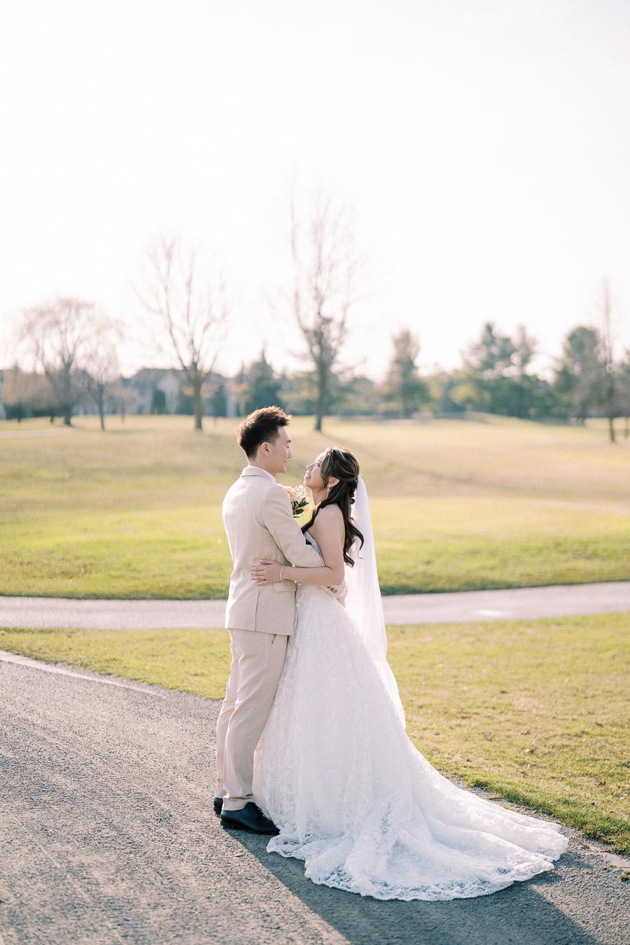 Romantic moment between a couple dressed in wedding attire on a sunlit lawn, with a bride in a flowing white gown and veil, embracing the groom in a light suit, set against a serene, natural landscape backdrop.