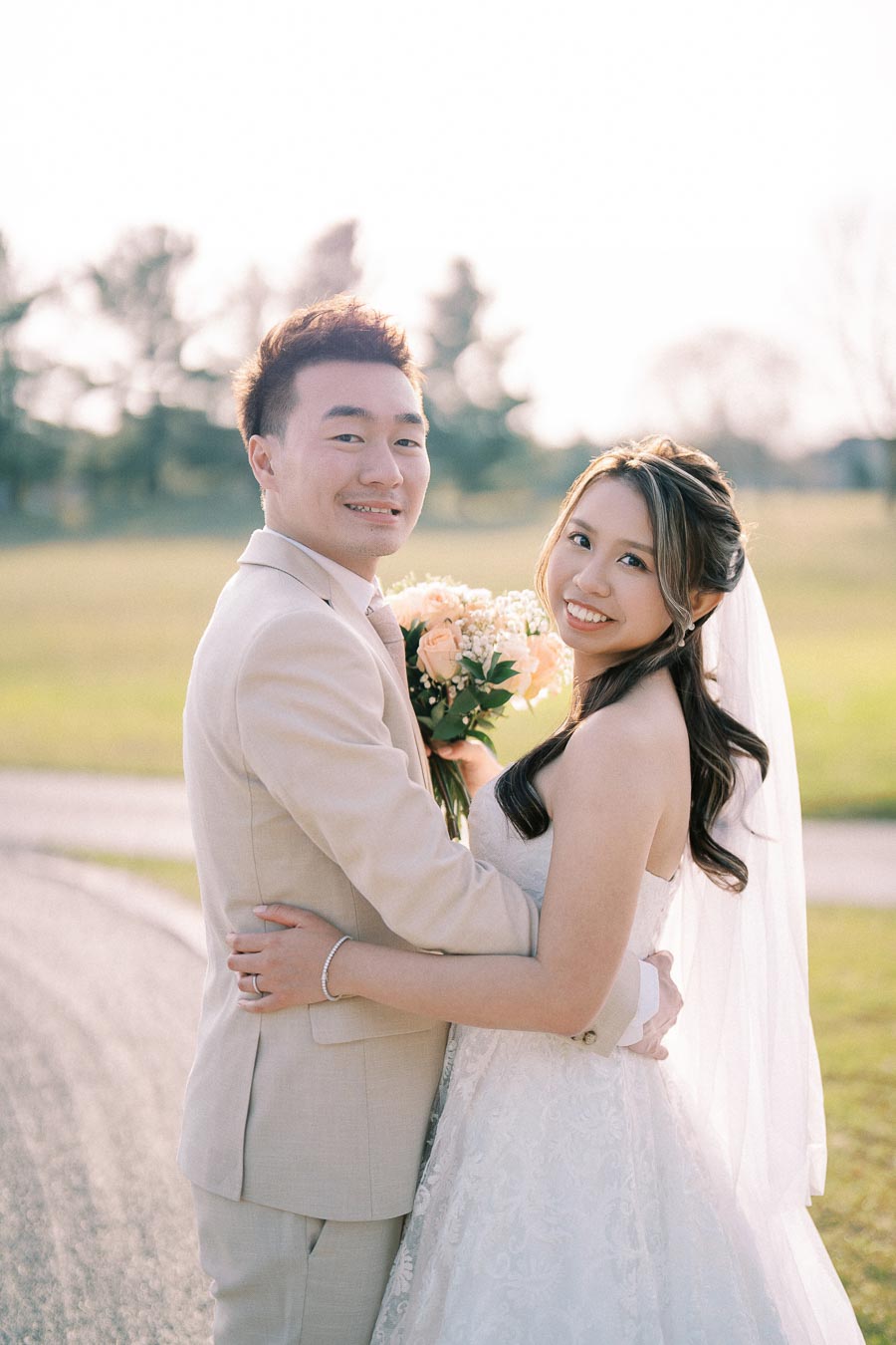 A happy couple embracing on their wedding day, with the bride in a white gown holding a bouquet of peach and white flowers, and the groom in a light beige suit, standing in a sunlit outdoor setting.