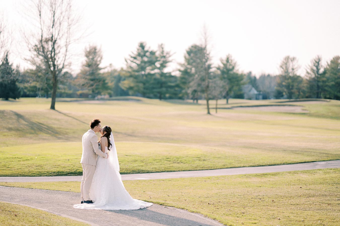 A bride and groom embrace and kiss on a picturesque golf course, surrounded by lush greenery and bare trees, under a clear sky, capturing a romantic outdoor wedding moment.