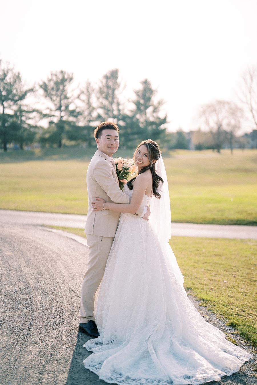 Elegant outdoor wedding photo of a smiling bride in a white lace gown and groom in a beige suit embracing in a sunlit park.