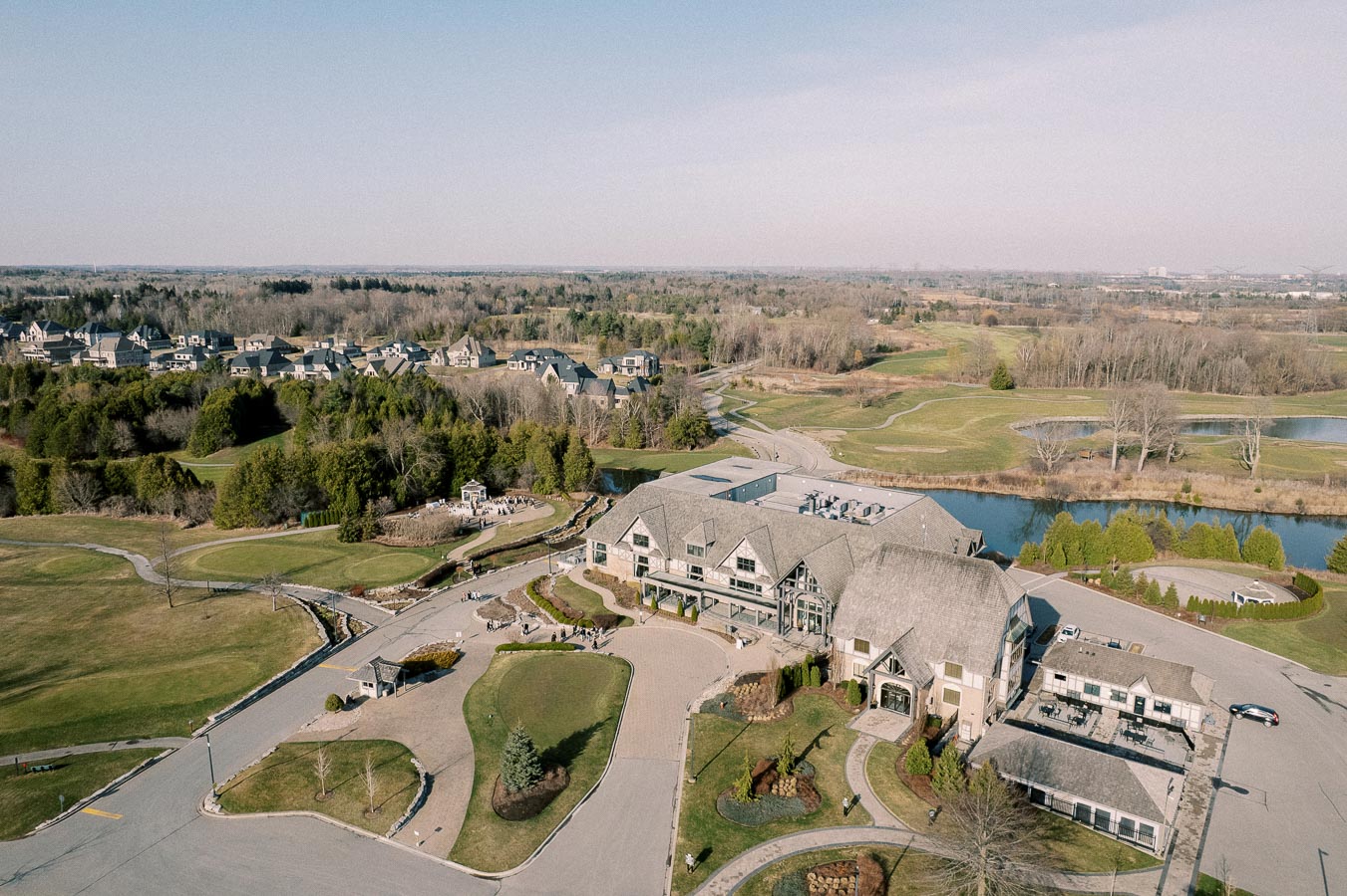 Aerial view of an expansive golf course with a clubhouse surrounded by lush greenery, residential homes, and a calm river, under a clear blue sky.