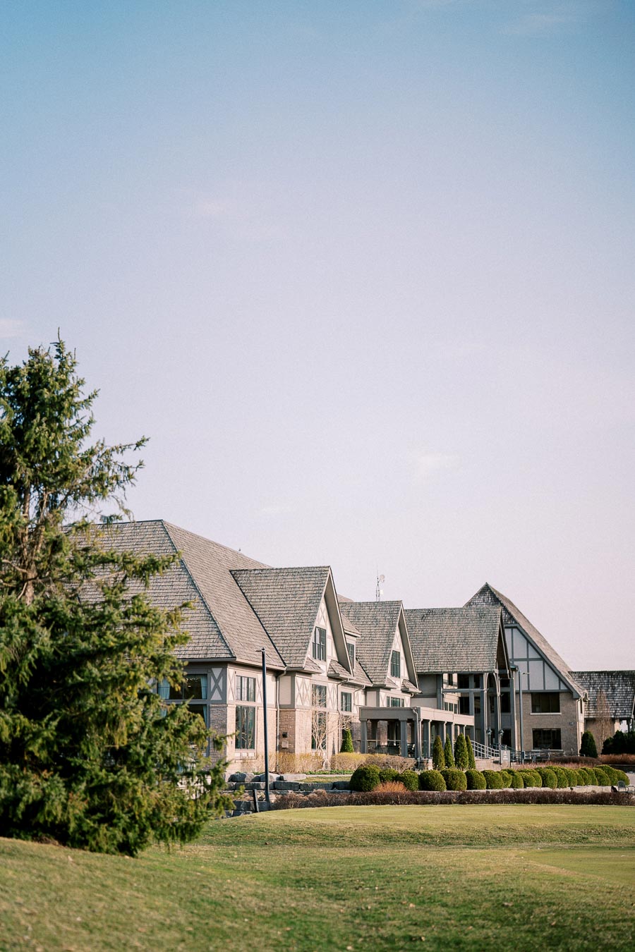 Elegant Tudor-style building surrounded by lush greenery, featuring a spacious lawn and clear blue sky background.
