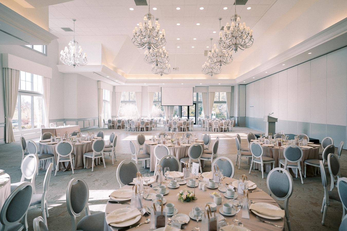 Elegant wedding venue with beautifully arranged banquet tables, chandeliers, and large windows allowing natural light to fill the spacious room.