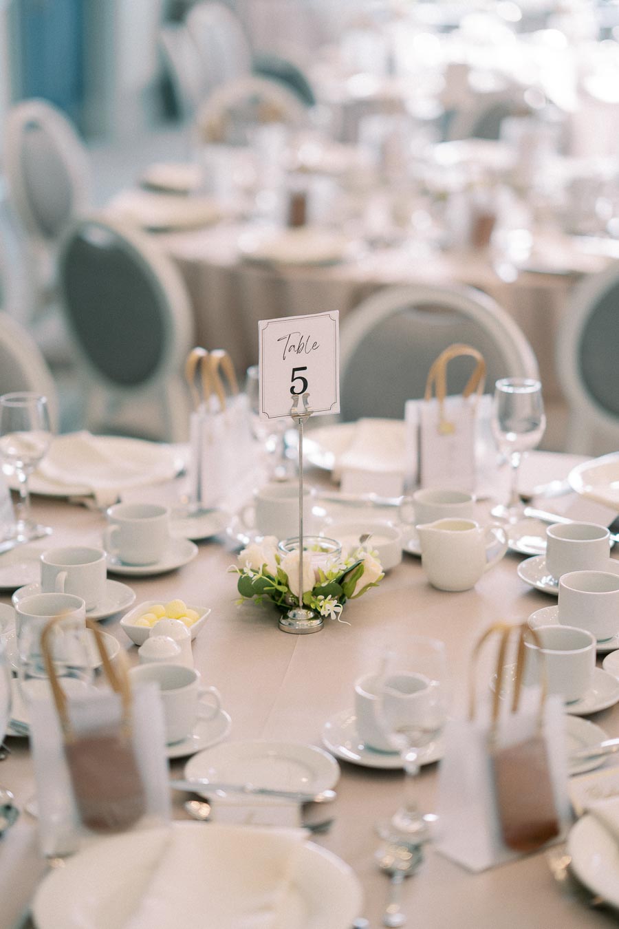 Elegant wedding reception table set-up with table number 5. The table is adorned with white dishes, teacups, and delicate floral centerpiece. Soft lighting enhances the sophisticated ambiance, ideal for a formal event.