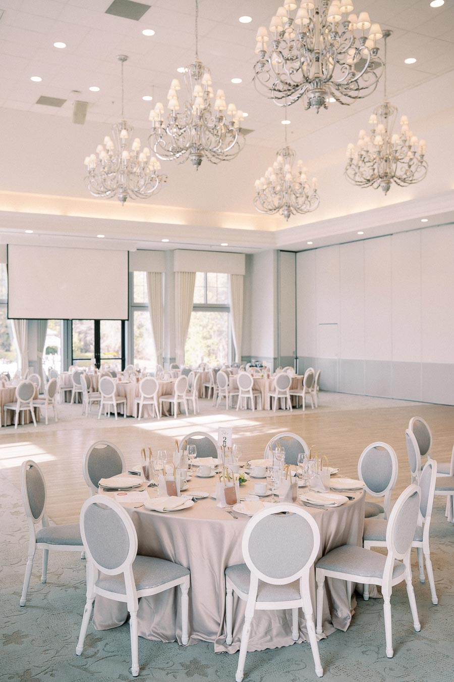 Elegant wedding reception venue with round tables set for dining, luxurious chandeliers, and natural light filtering through large windows.