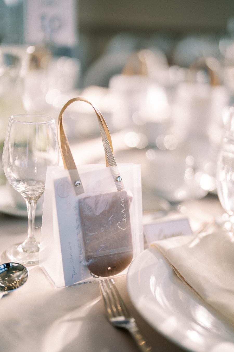 Elegant wedding table setting with a personalized gift bag holding a card and chocolate, placed next to a wine glass and a neatly folded napkin on a white plate, creating a sophisticated and inviting atmosphere.