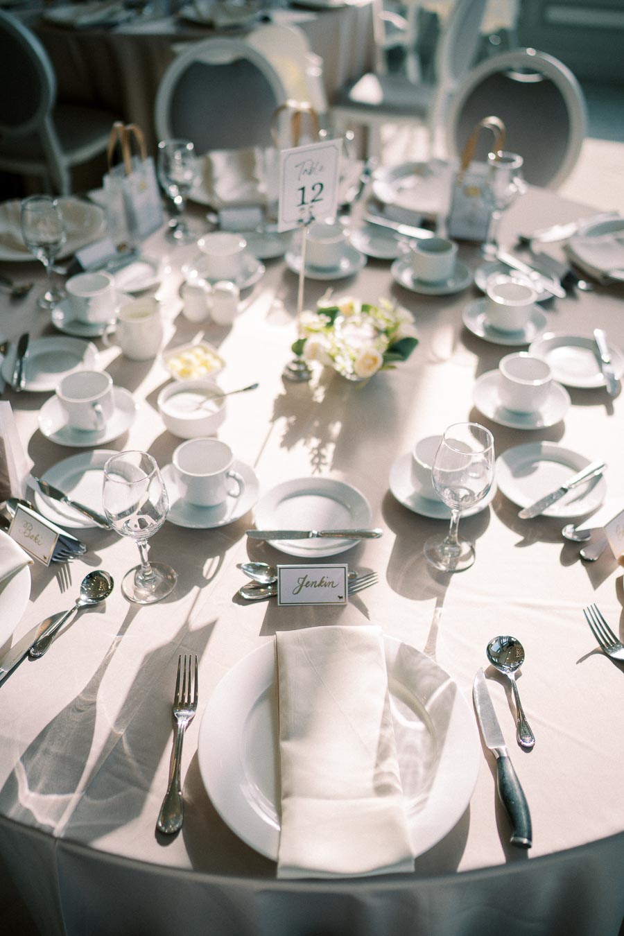 Elegant wedding reception table setting with white tableware, glassware, and a name card labeled Jenkin, surrounded by soft ambient lighting and floral centerpiece at Table 12.