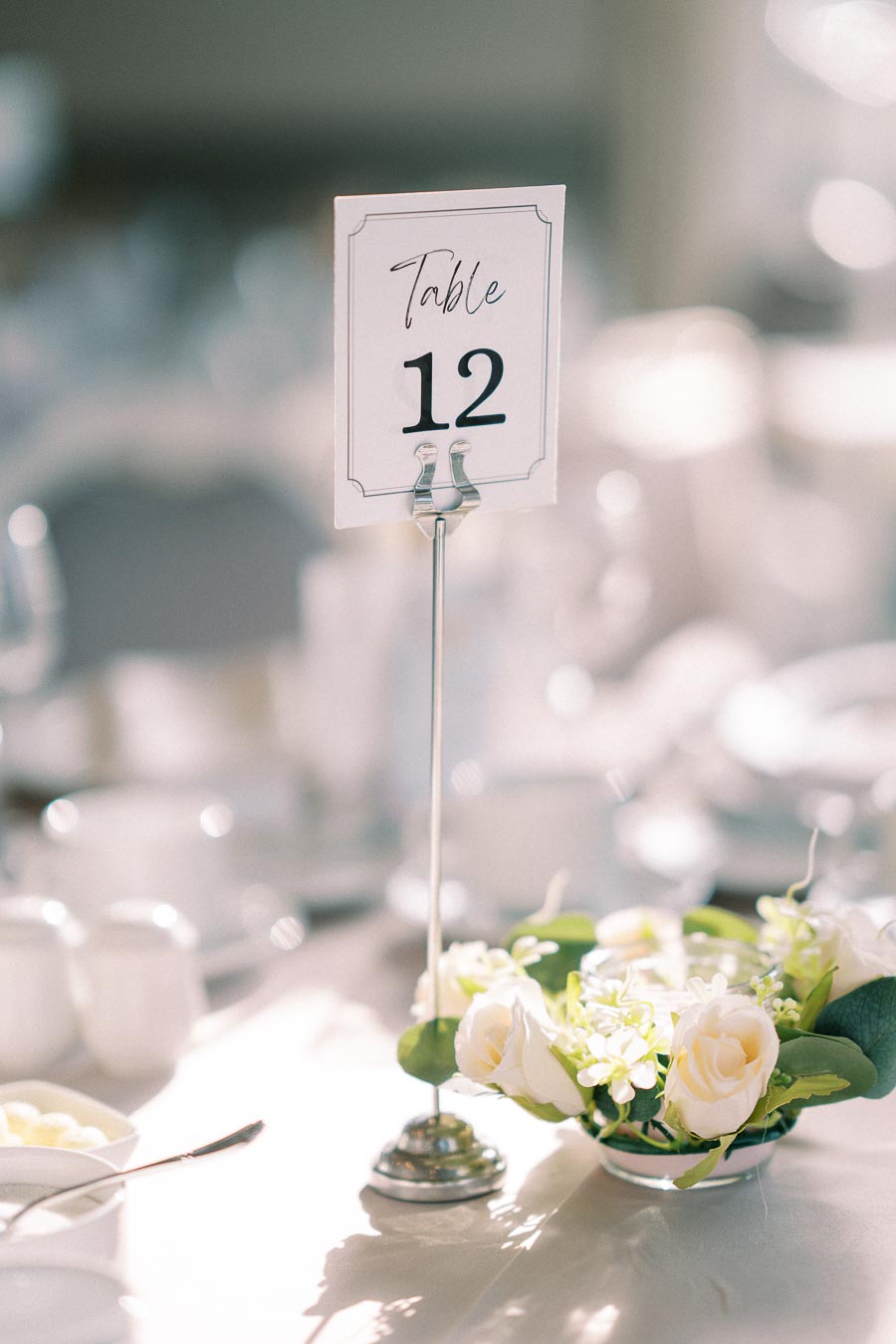 Elegant table setting with a Table 12 card on a stand, surrounded by white roses and greenery, at a wedding or formal event.
