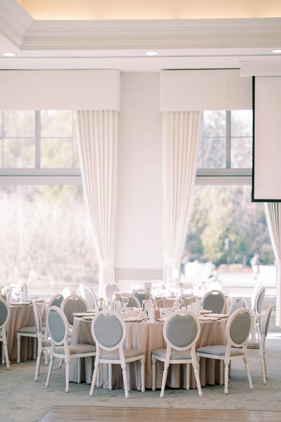 Elegant wedding reception hall with round tables, set with fine china and crystal glassware, surrounded by plush chairs and framed by large windows with white curtains.