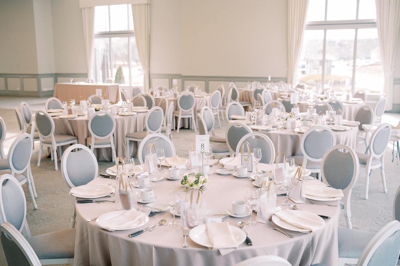 Elegant banquet hall set for a formal event with round tables, white tablecloths, fine china, glassware, and gray cushioned chairs, large windows providing natural light.