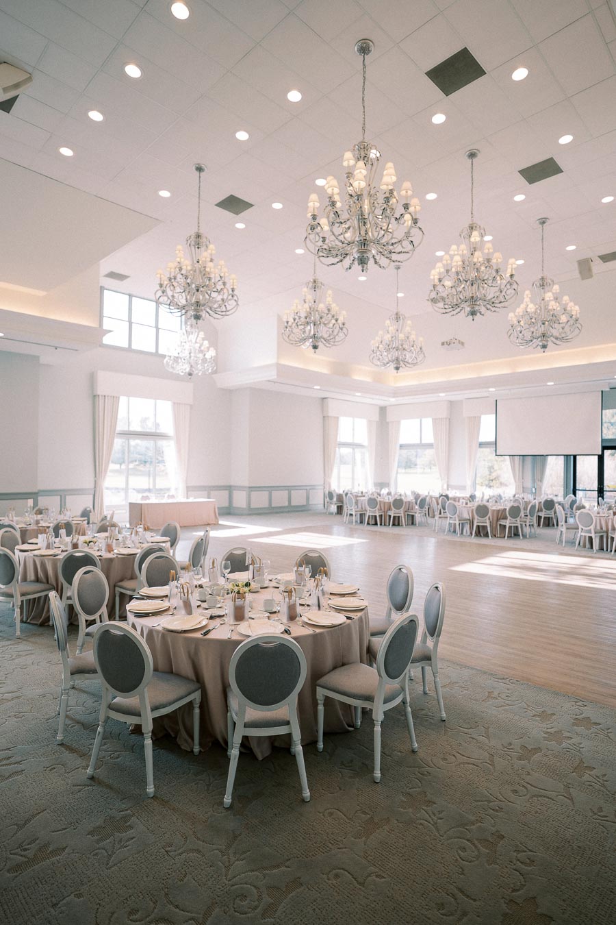 Elegant banquet hall with round tables set for a formal event, featuring luxurious chandeliers and large windows allowing abundant natural light.