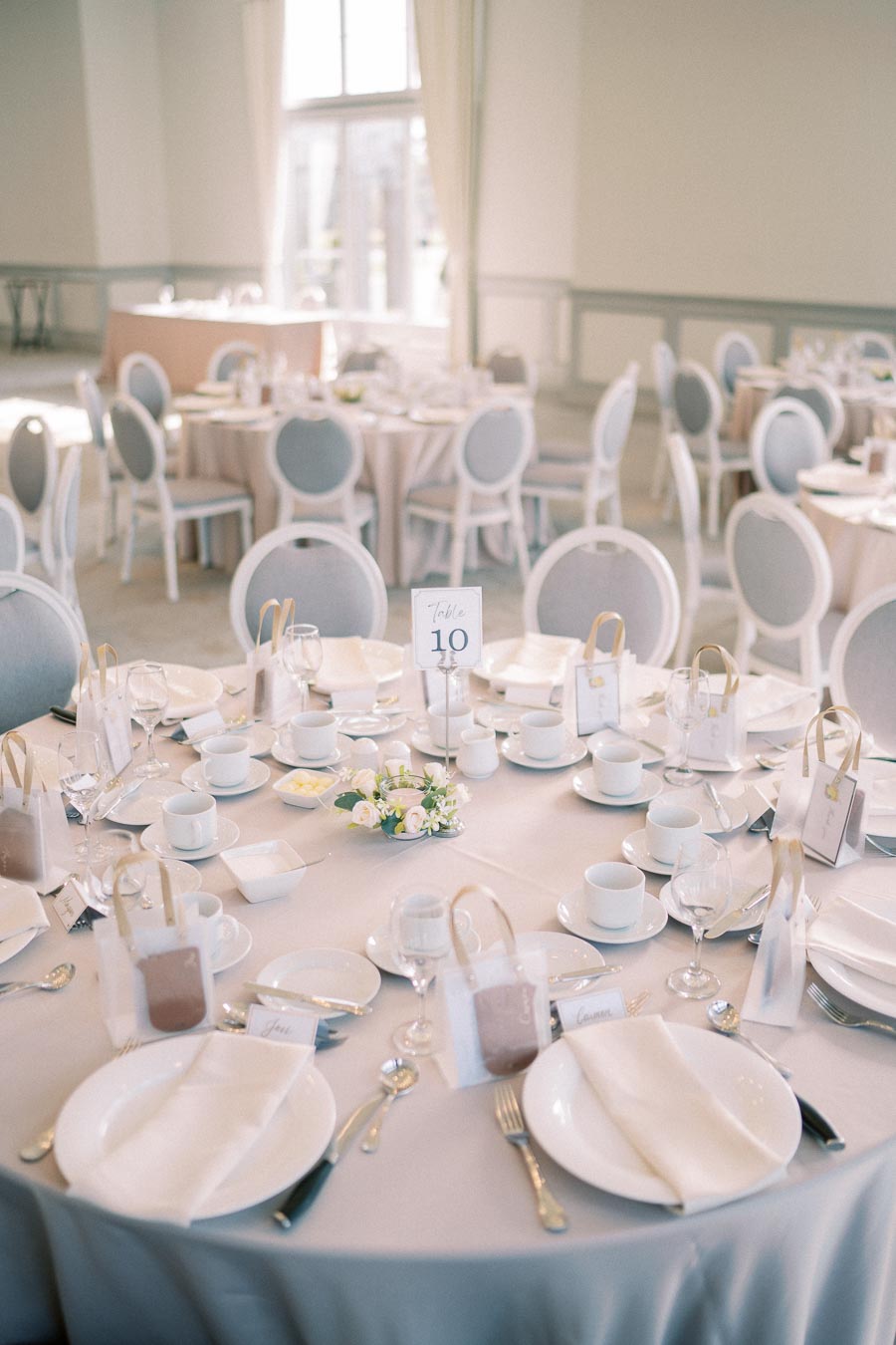 Elegant wedding reception table setting with white plates, cutlery, and gift bags on a pastel tablecloth. Featuring delicate floral centerpieces and a table number sign, in a bright, well-lit venue with soft-hued chairs and decor.