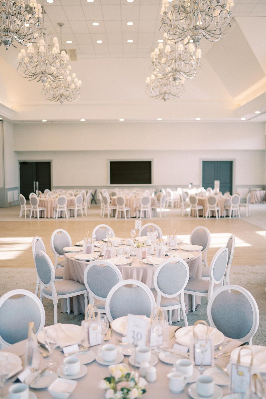 Elegant wedding reception hall with round tables set for dining, featuring white tablecloths, neatly arranged plates and glasses, and decorative chandeliers hanging from the ceiling.