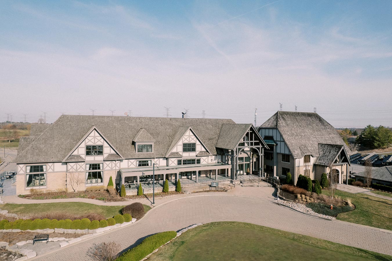 Aerial view of a large, Tudor-style clubhouse with a stone and wood facade, surrounded by landscaped gardens and paved walkways under a clear blue sky.