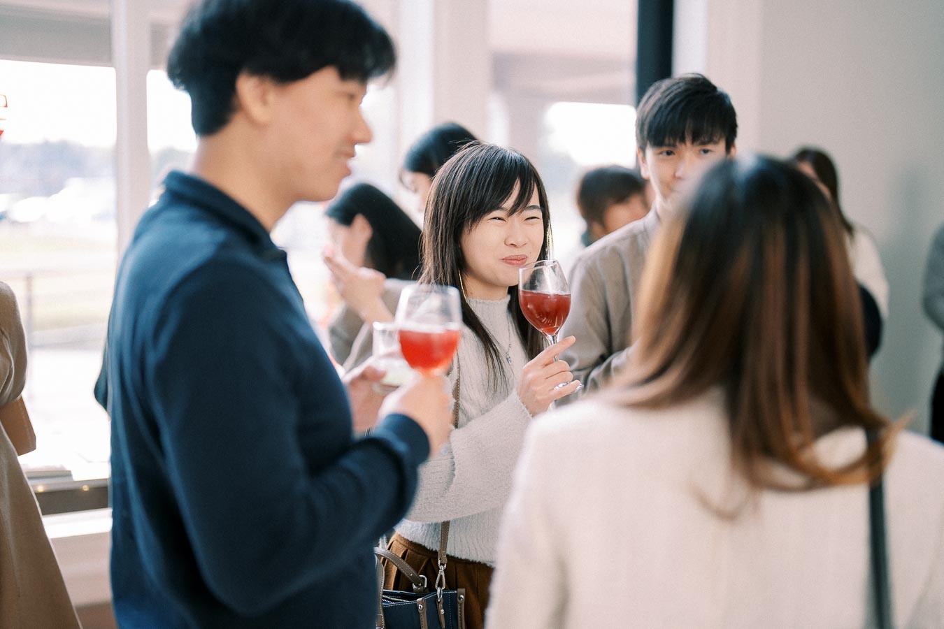 A group of people enjoying drinks at a social gathering, focusing on a woman smiling with a glass of red beverage in hand.