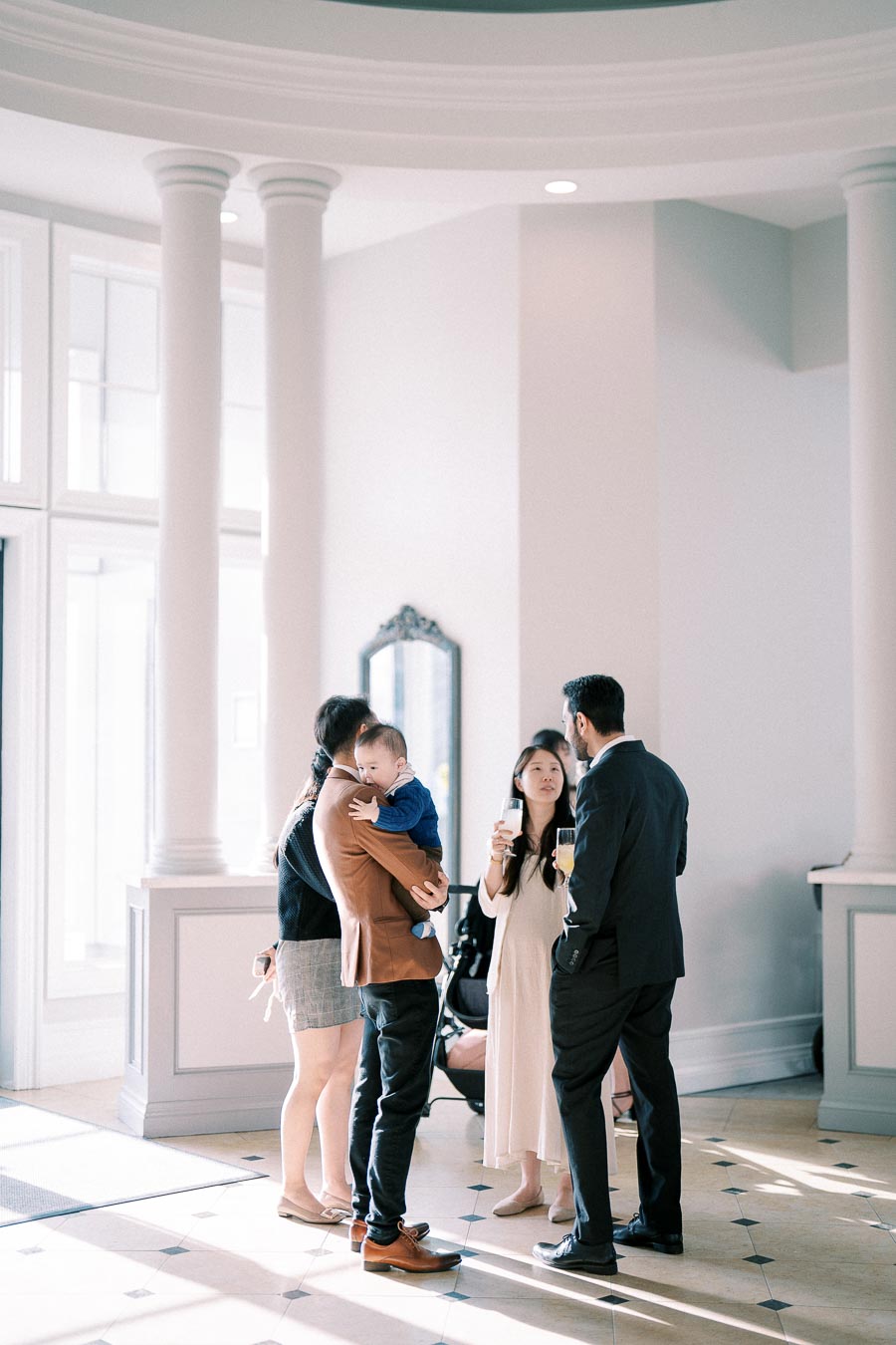 A small group of well-dressed people conversing in a bright, elegant room, with one individual holding a baby.