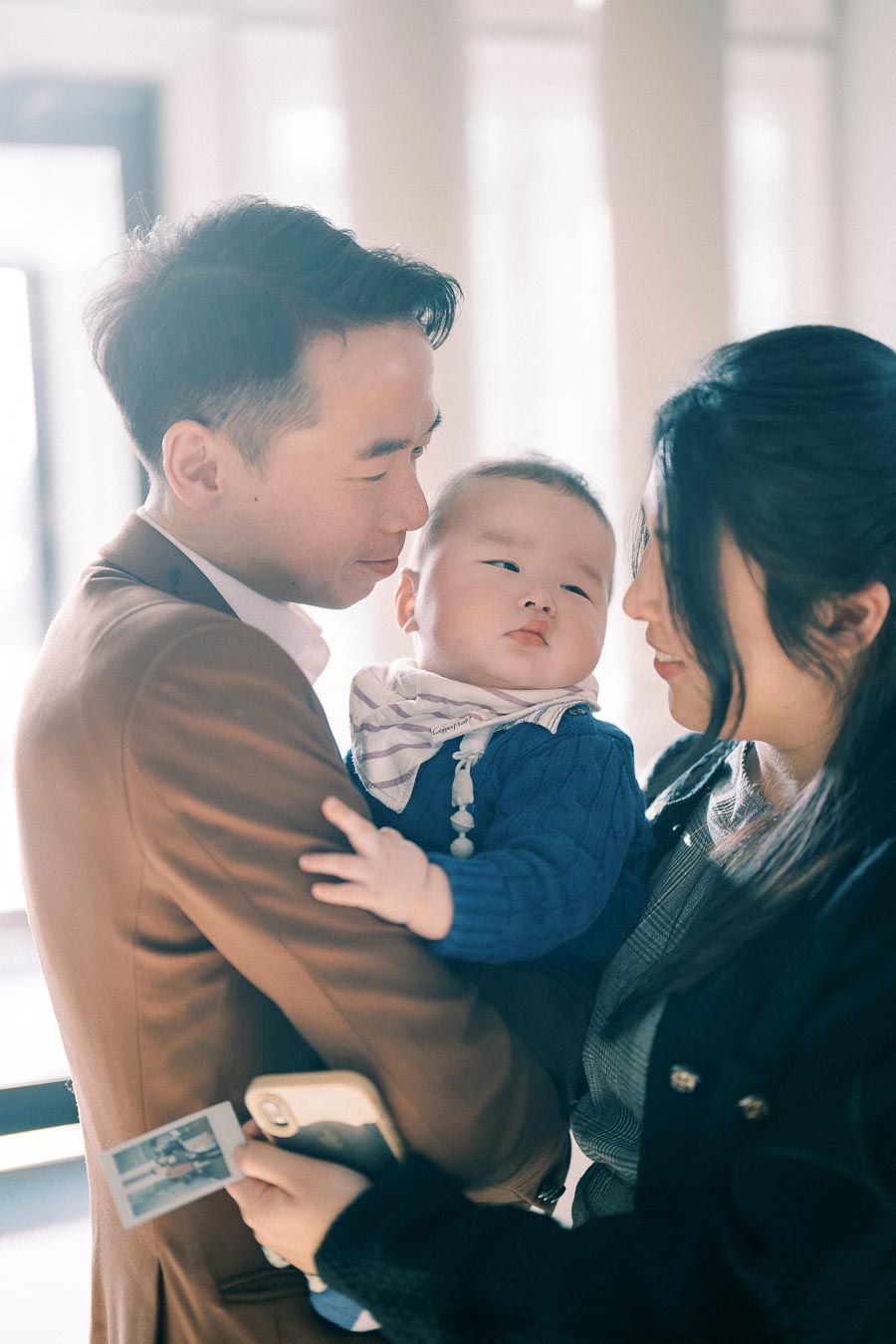A loving couple holds their baby, capturing a heartwarming family moment indoors, with soft natural lighting. The father, in a brown blazer, smiles affectionately at the baby, while the mother looks on tenderly, holding a smartphone and a small photo.