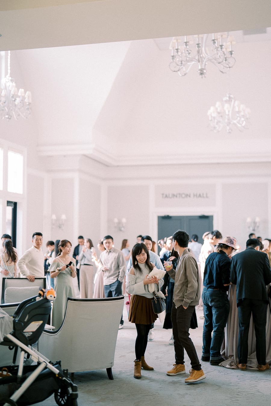 A lively social gathering taking place at Taunton Hall, featuring a diverse group of people engaging in conversation and enjoying refreshments in a bright, elegant setting with chandeliers and natural light.