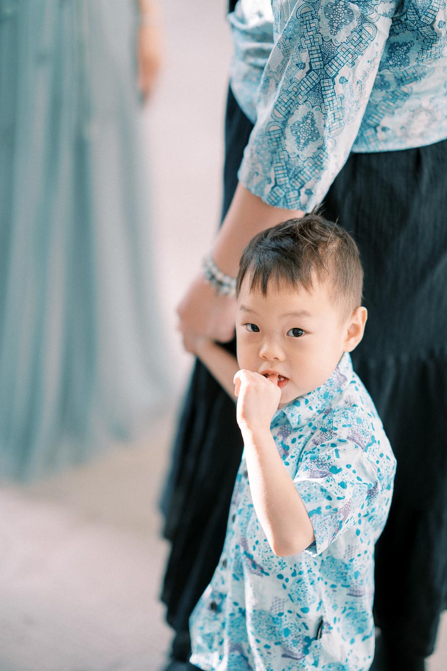 Child holding an adult's hand, wearing a light blue patterned shirt, looking towards the camera with a curious expression.