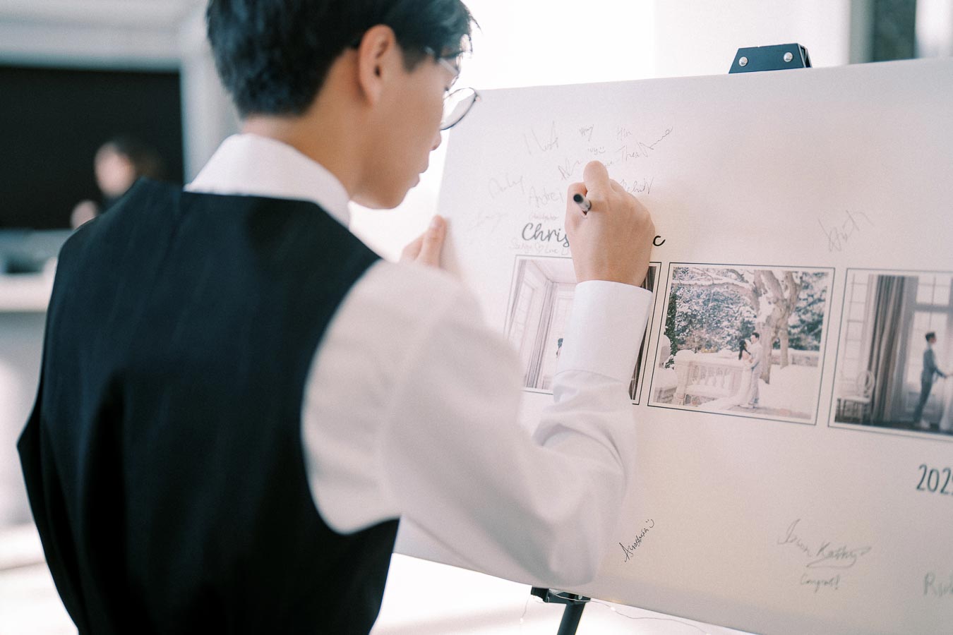 A person in formal attire signing a wedding guest book with photos displayed, capturing an elegant and memorable moment.