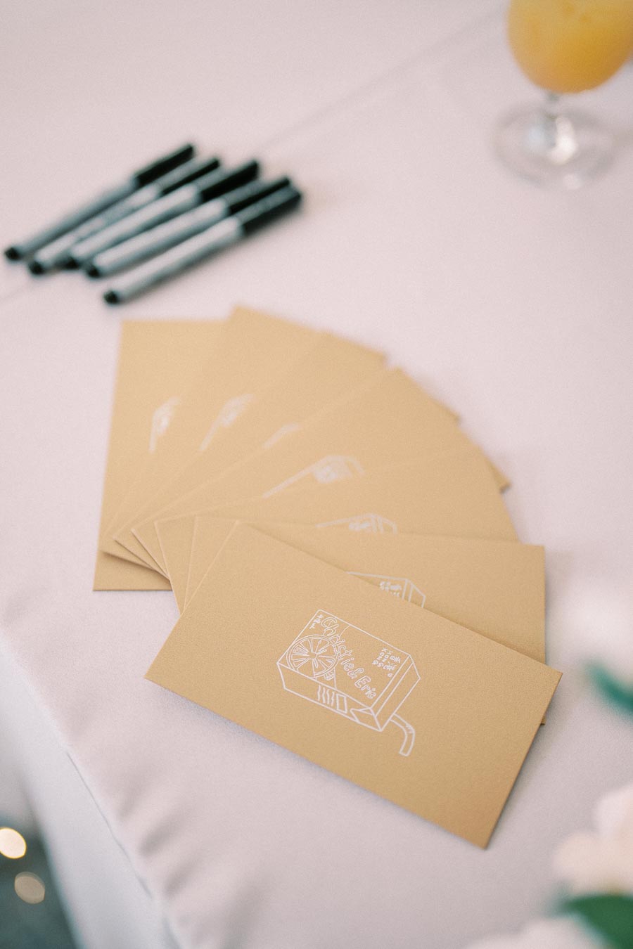 Stack of beige envelopes with white illustrations on a table, alongside several black pens and a glass of orange juice in the background.