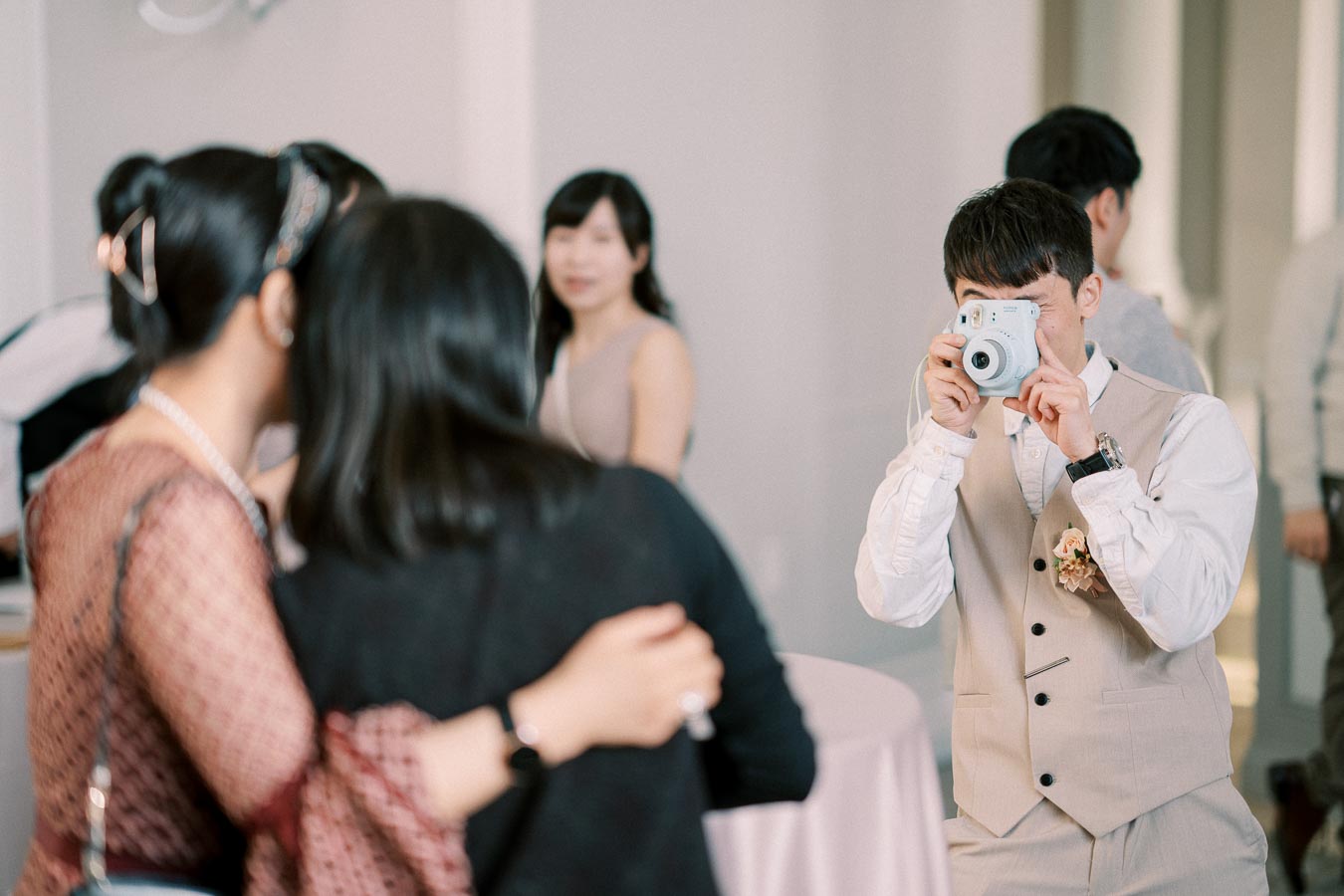 A young man in a beige suit taking a photo with an instant camera at a social event, while two women in the foreground embrace and pose, with other guests in the background.