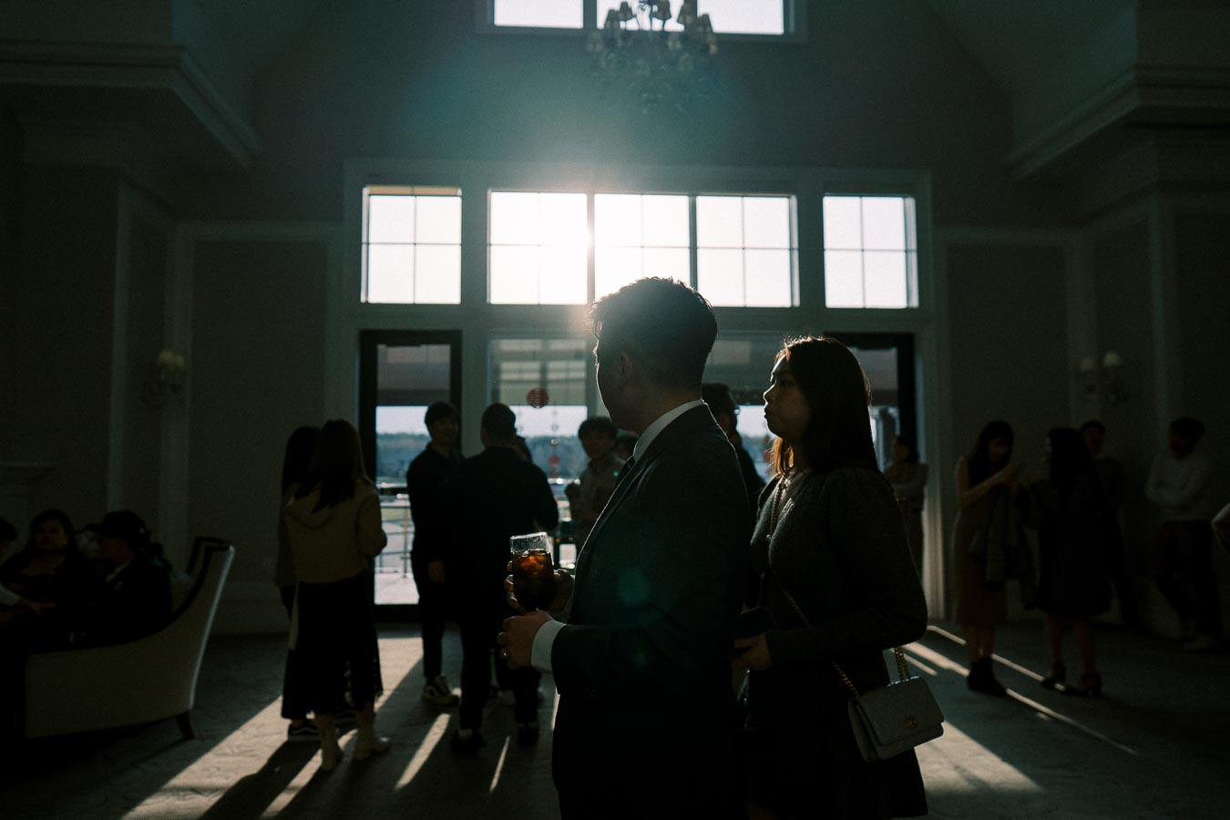 A group of people at an indoor social gathering, with sunlight streaming through large windows, creating silhouettes and a warm atmosphere.