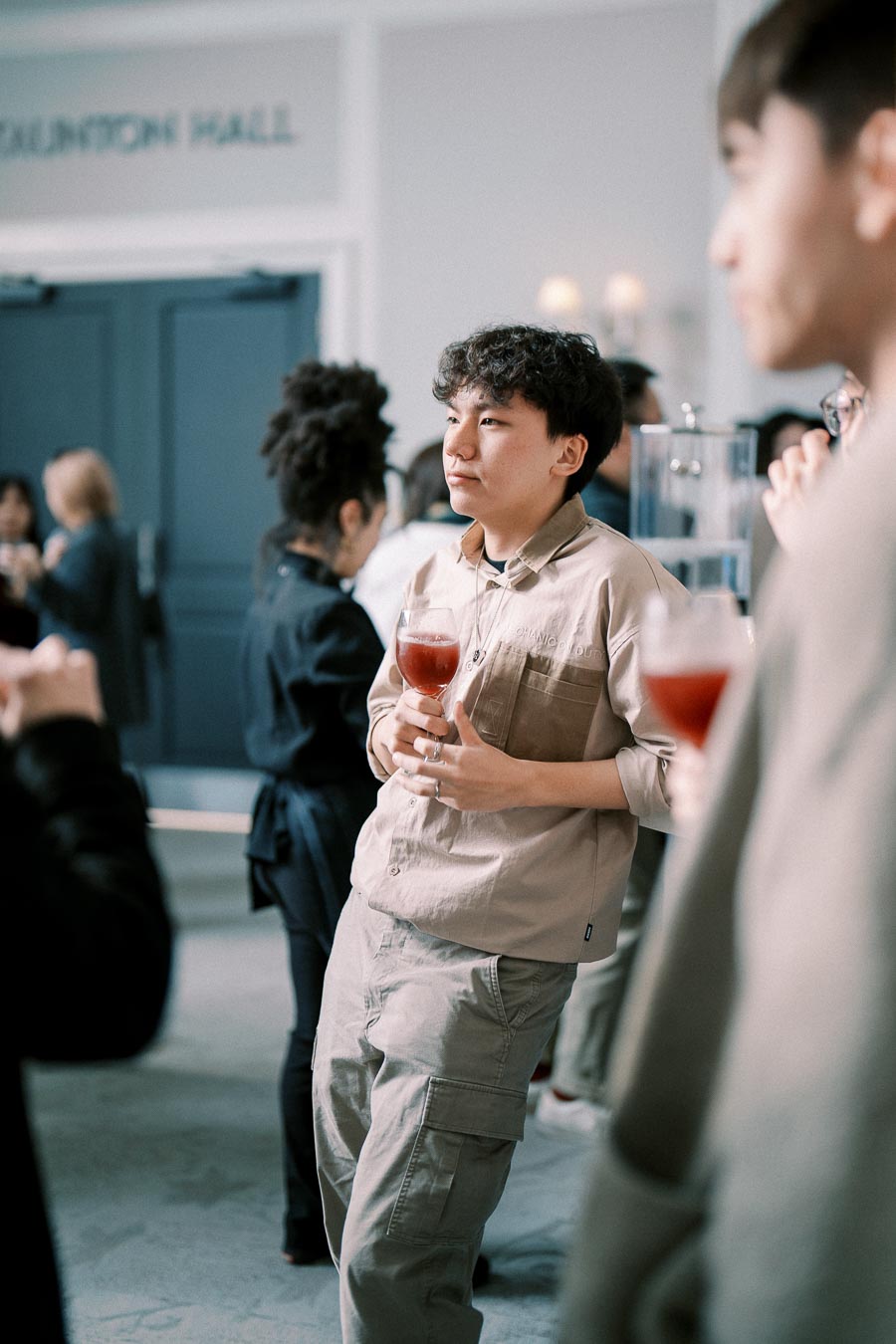 Young man at a social gathering holding a red drink, standing in a relaxed pose with other attendees in a modern event hall.