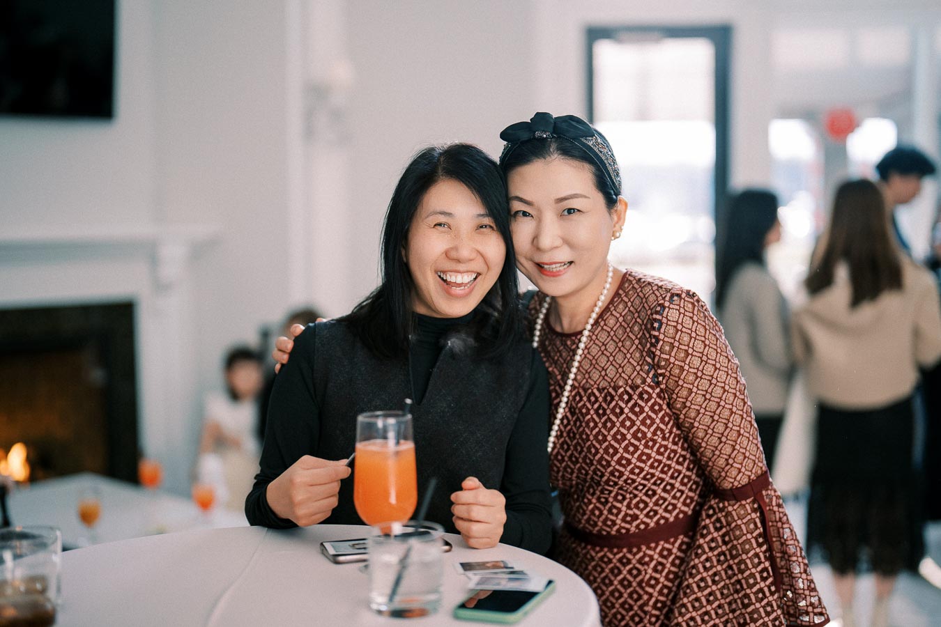 Two women smiling and posing together at a social gathering, with one holding an orange drink in a glass. The background shows a cozy indoor setting with other people mingling.