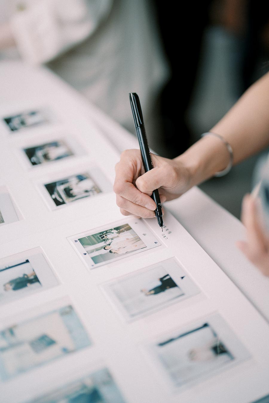 A person's hand writing notes next to a series of instant photos on a white table, possibly for a photo album or memory board project.