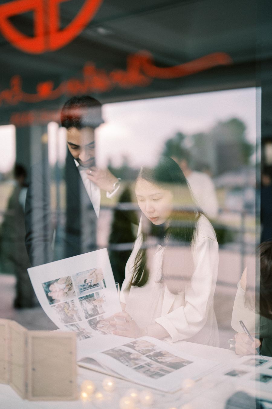Two people reviewing a photo album through a glass window, with soft reflections and indoor ambient lighting.