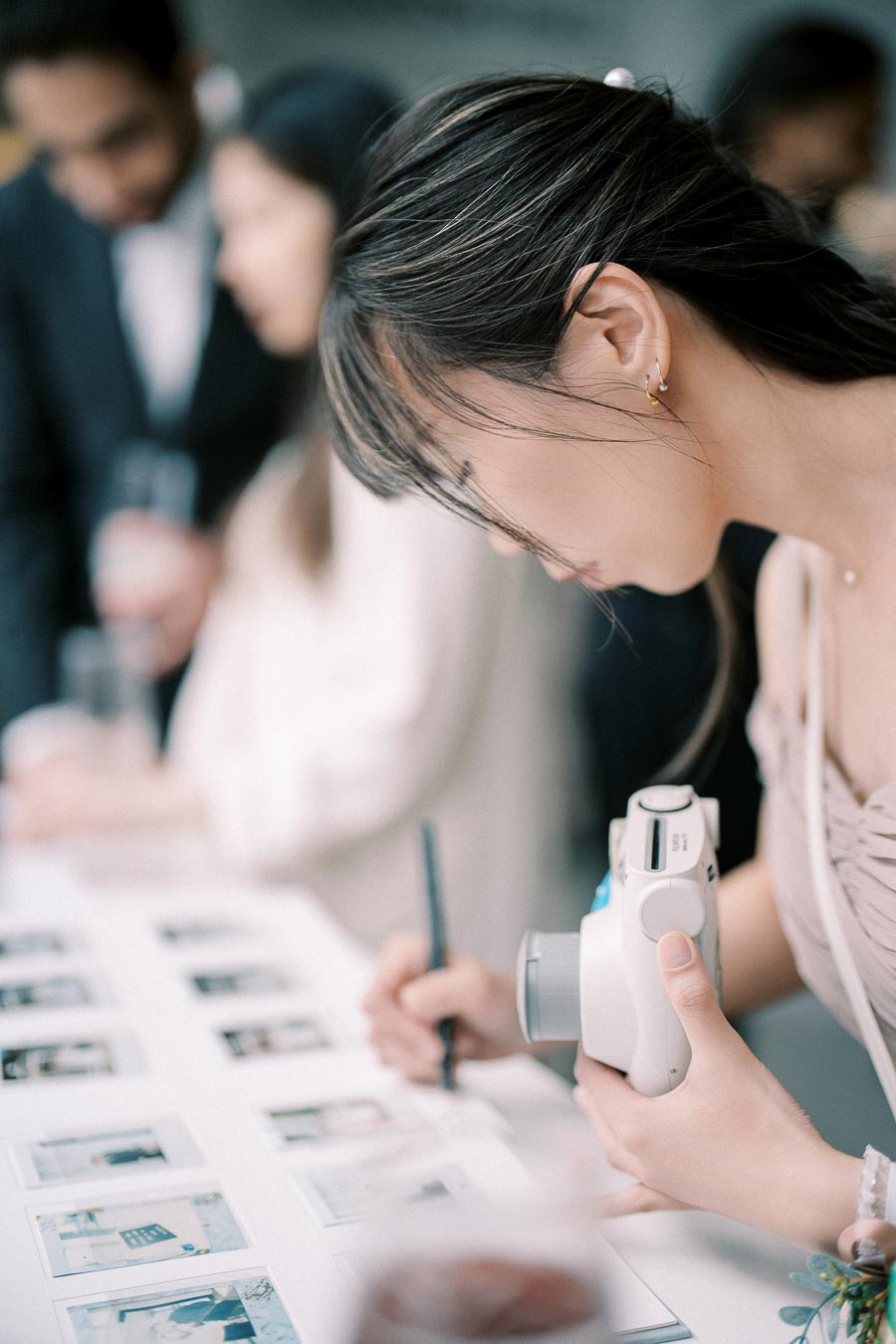 Young woman signing guest book with polaroid camera in hand at a formal event, blurred attendees in background.