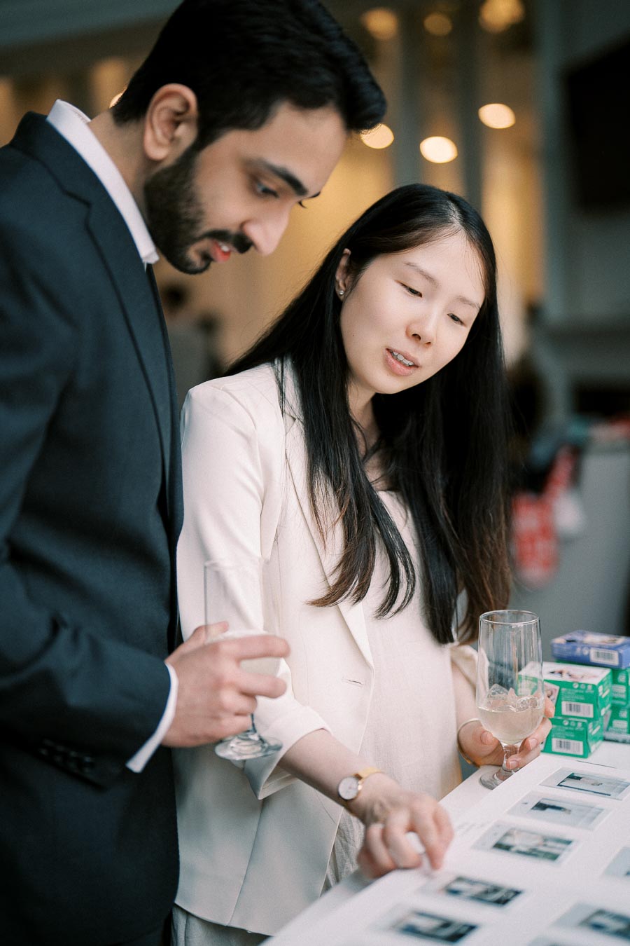Two professionals in formal attire reviewing photographs on a table while holding wine glasses, with a focused and collaborative interaction.