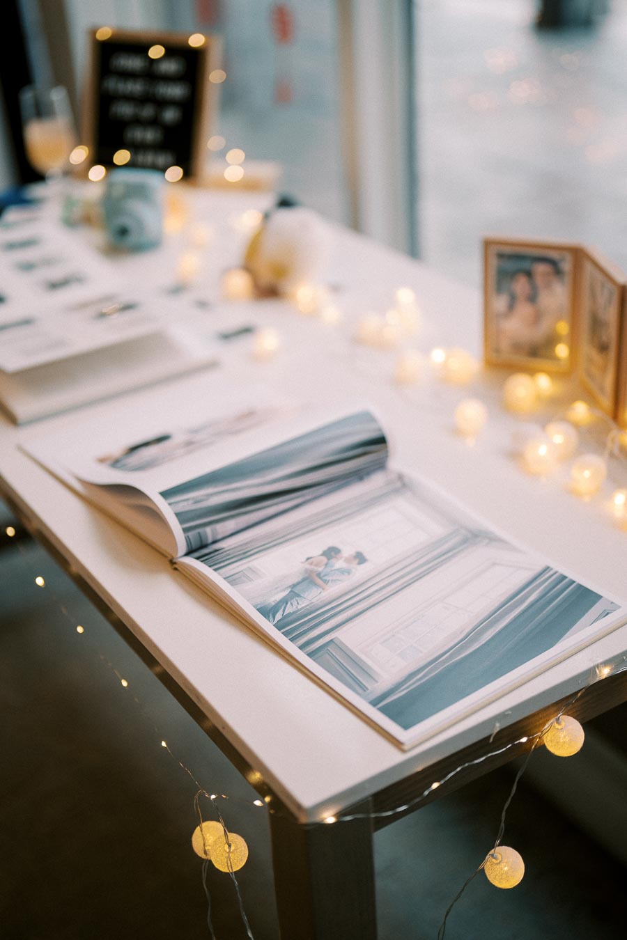 A beautifully arranged wedding reception table featuring a photo album with a romantic couple image, surrounded by soft, glowing string lights, elegant photo frames, and a stylish camera, creating a warm and intimate ambiance.