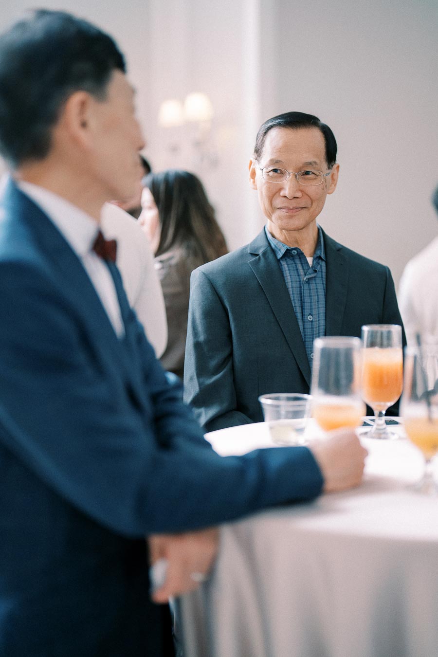 A man in a suit standing at a table with drinks during a social gathering, engaged in conversation with another person in a formal setting.