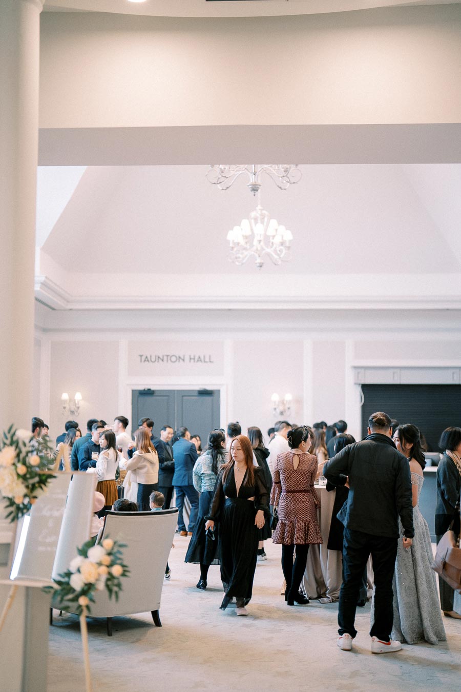 People mingling at a social event inside Taunton Hall, elegantly decorated with chandeliers and floral arrangements, creating a sophisticated atmosphere.