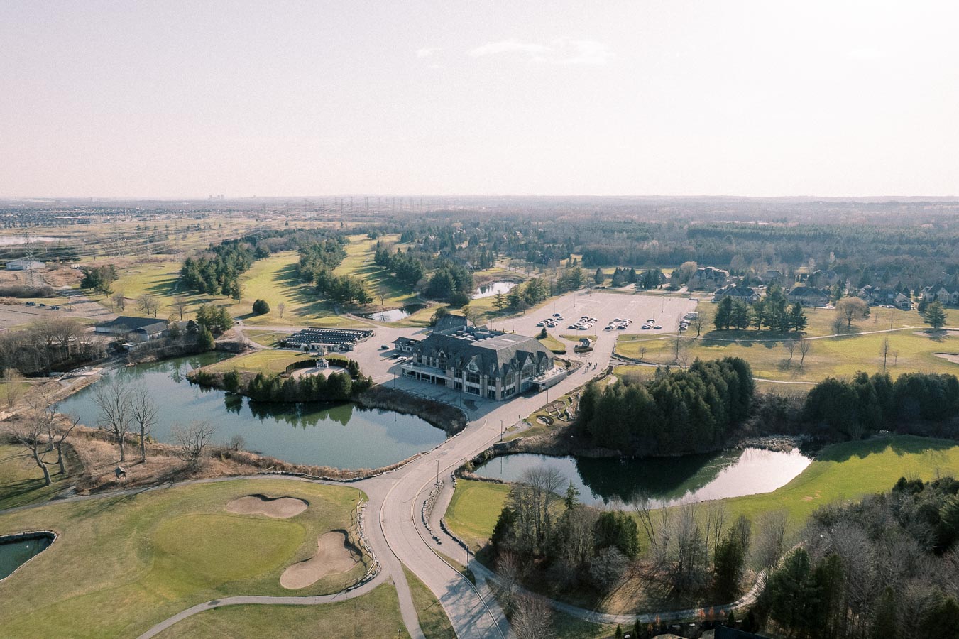 Aerial view of a scenic golf course surrounded by lush greenery and water features, featuring a clubhouse and nearby parking area under a clear sky.
