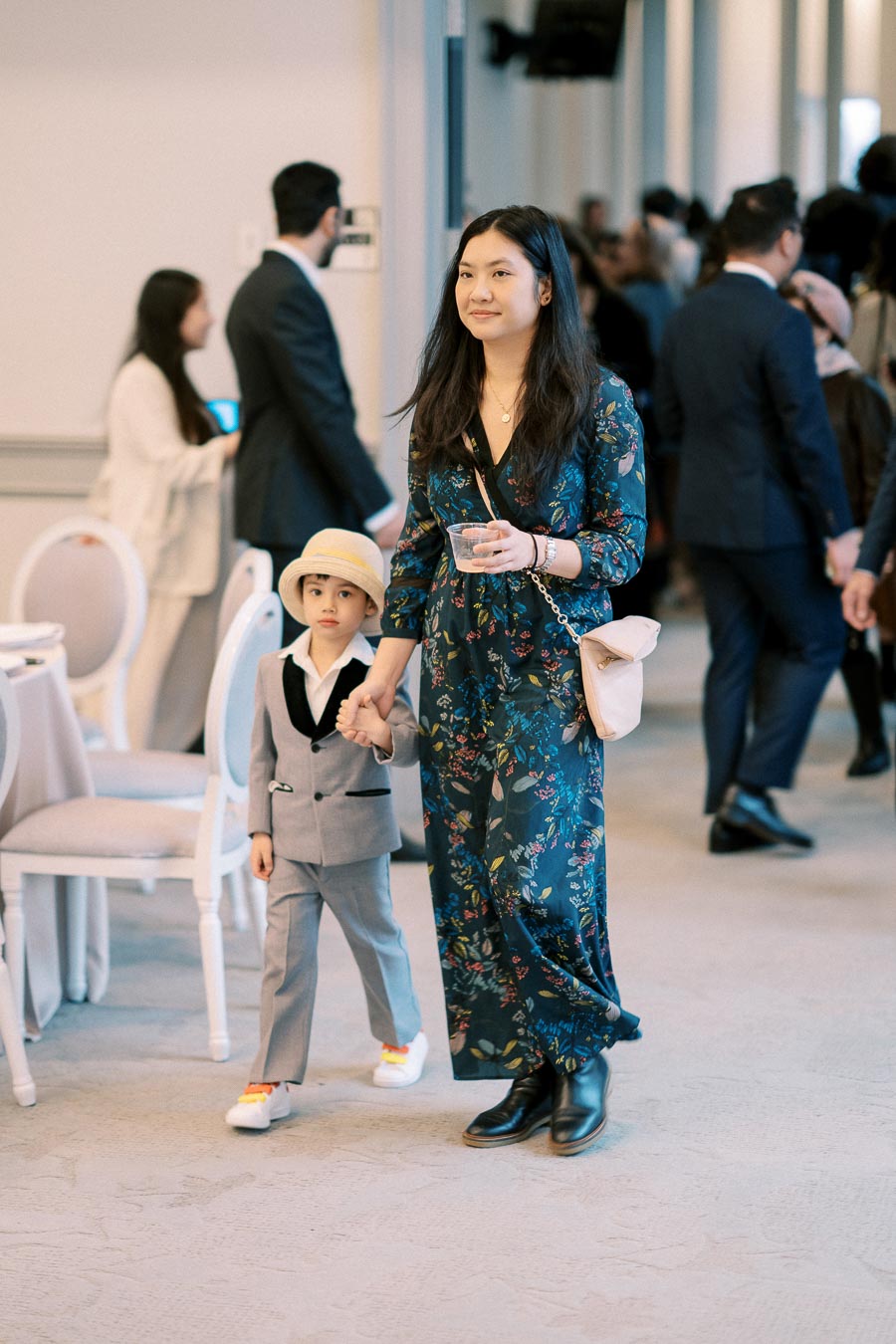 A woman in a floral dress holds hands with a young boy in a suit and hat at a formal event. The background shows several people in formal attire, creating a sophisticated atmosphere.