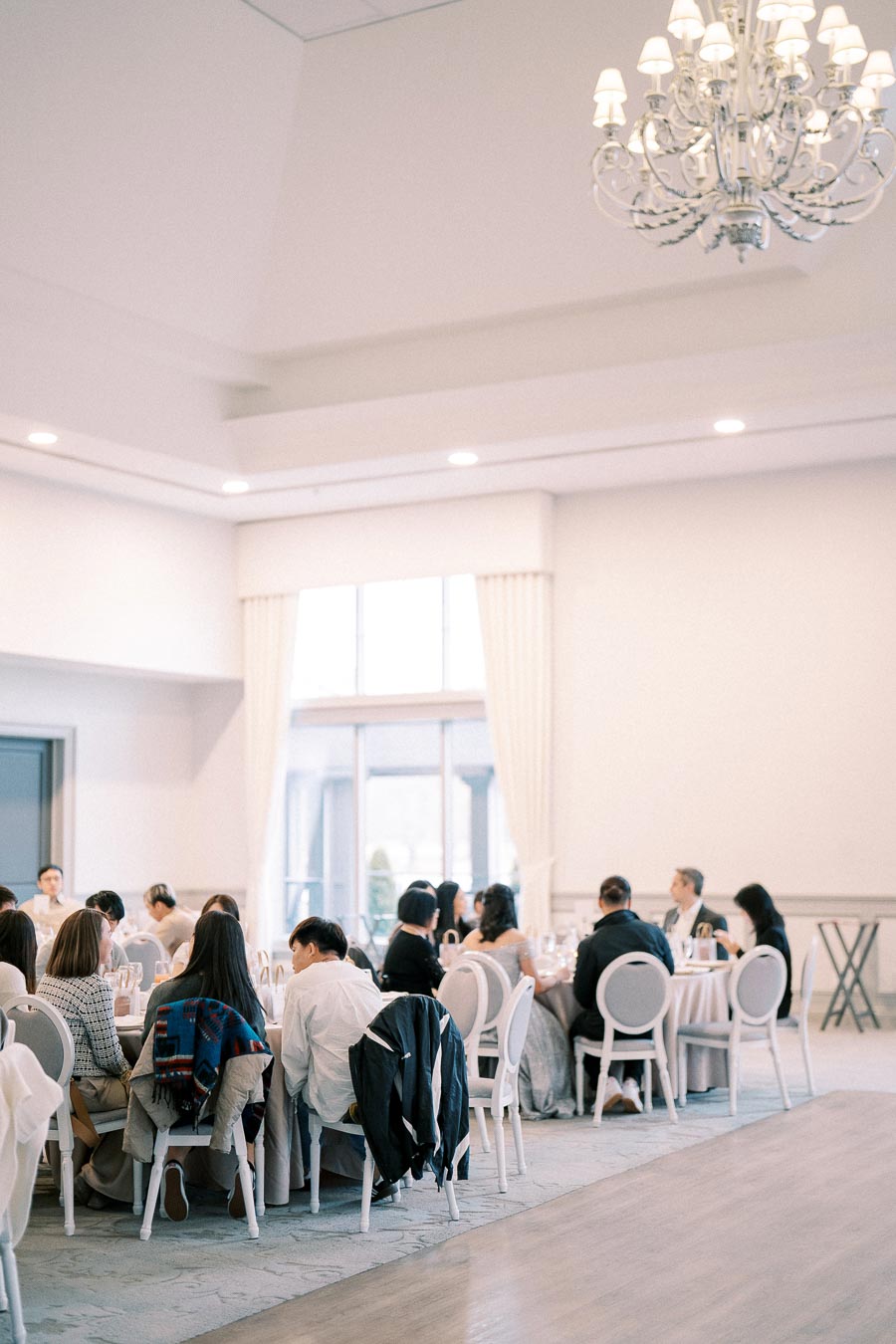 A group of people seated at round tables in an elegant banquet hall with a chandelier and tall windows, attending a formal event or reception.