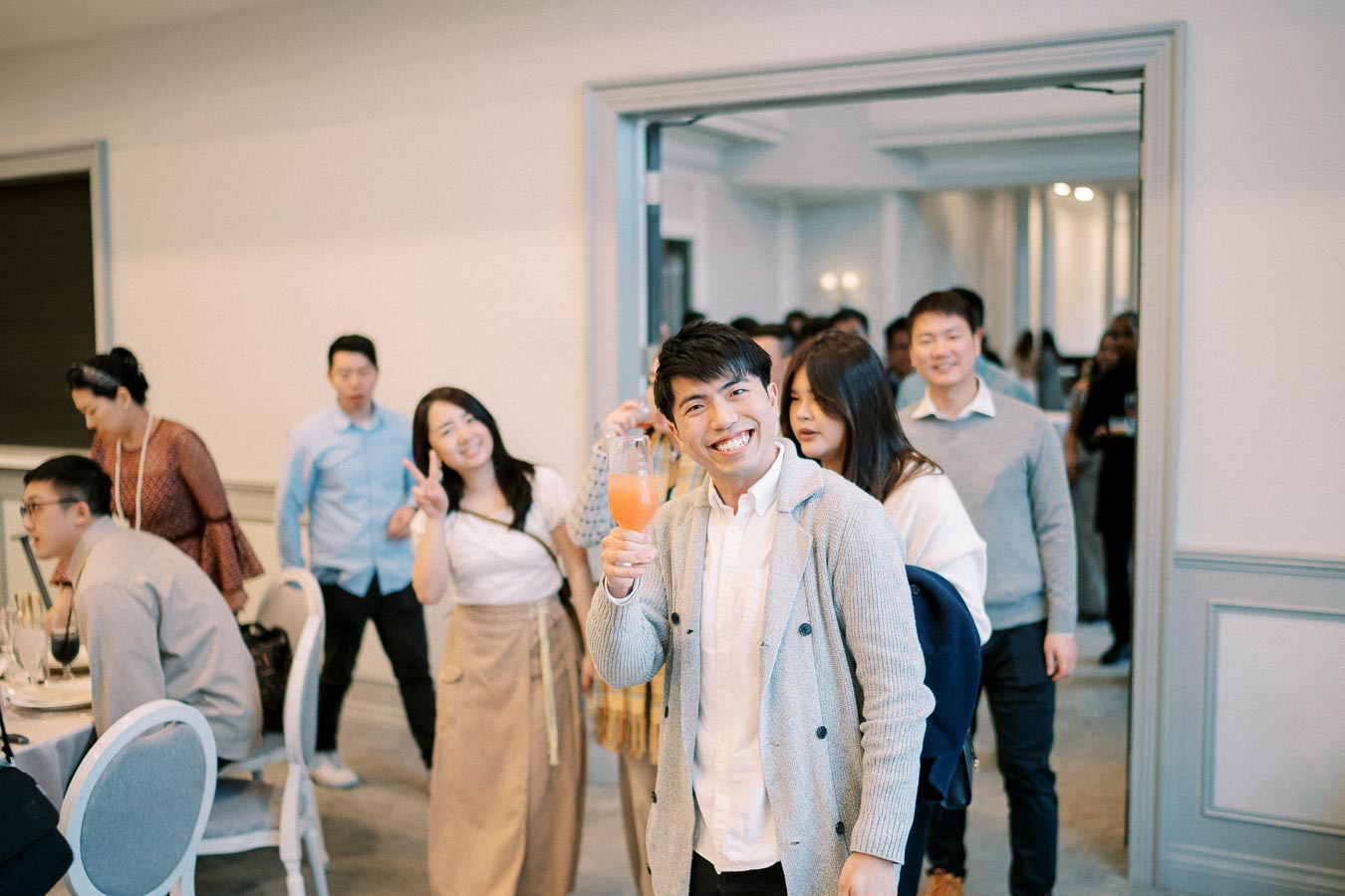 A cheerful group of people at a social gathering, with a man in the foreground smiling and holding an orange drink, surrounded by others enjoying the event indoors.