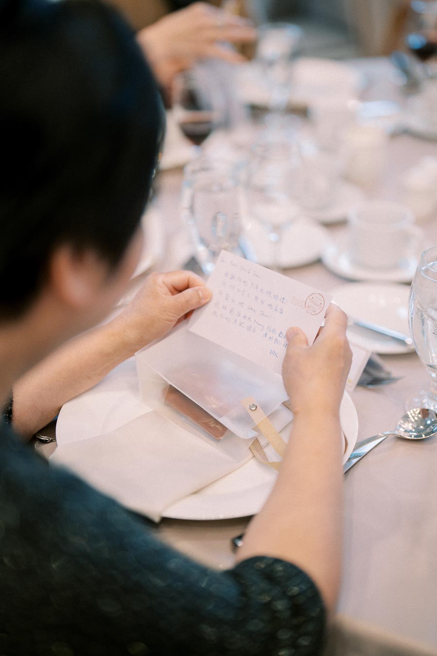 A person sitting at a dining table reading a card in a translucent envelope, surrounded by glassware and tableware.