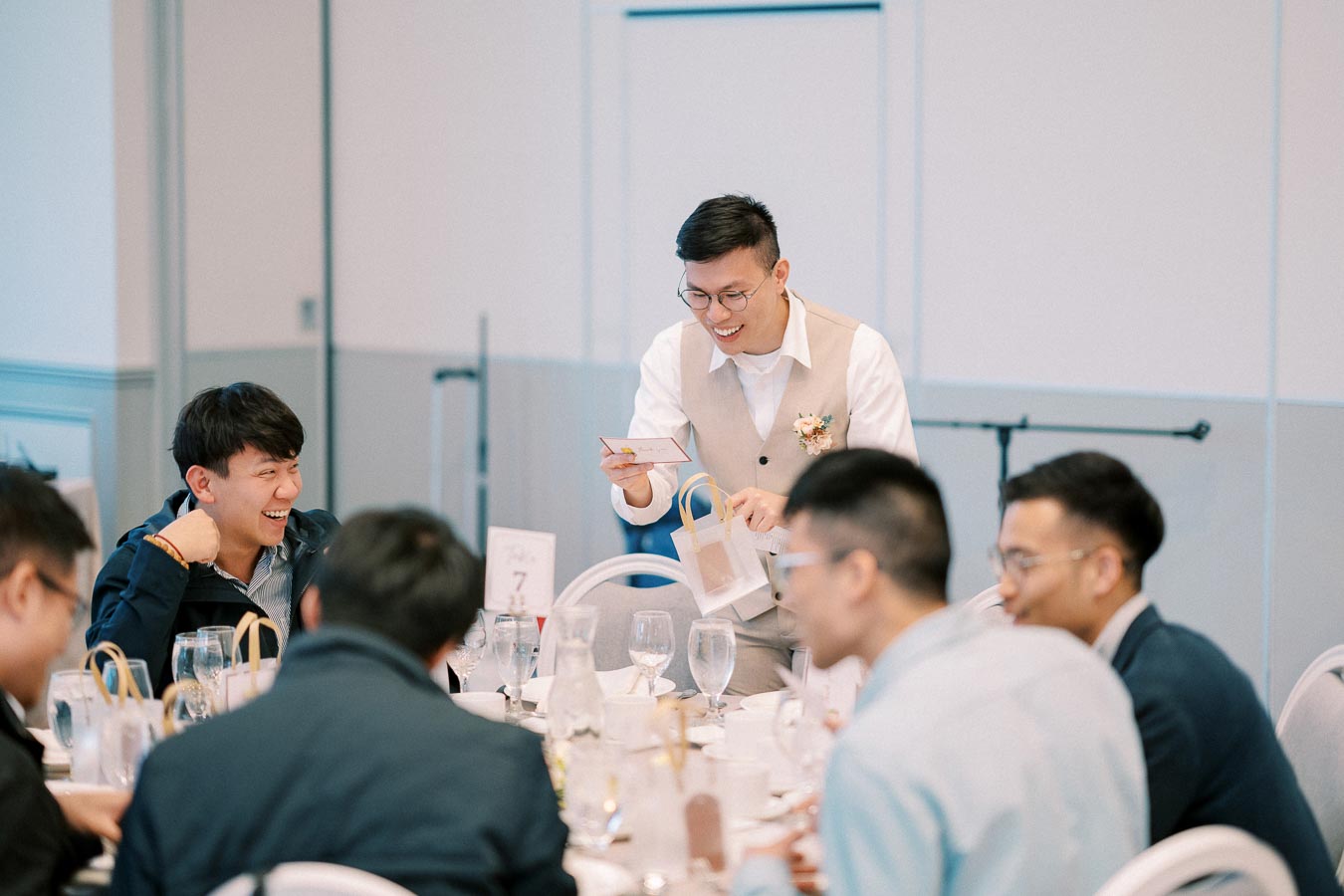 A group of men enjoying a joyful moment at a formal event, with one standing and handing out a small gift bag at a restaurant table, creating a lively and festive atmosphere.