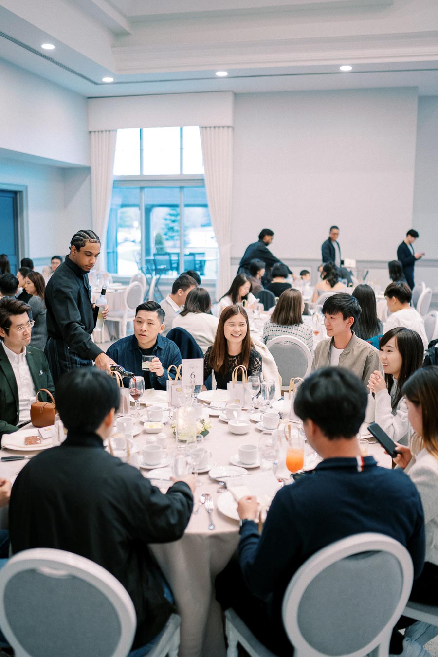 Group of people enjoying a formal dining event, seated around a round table set with place settings, glasses, and floral centerpieces, in a bright and spacious venue.