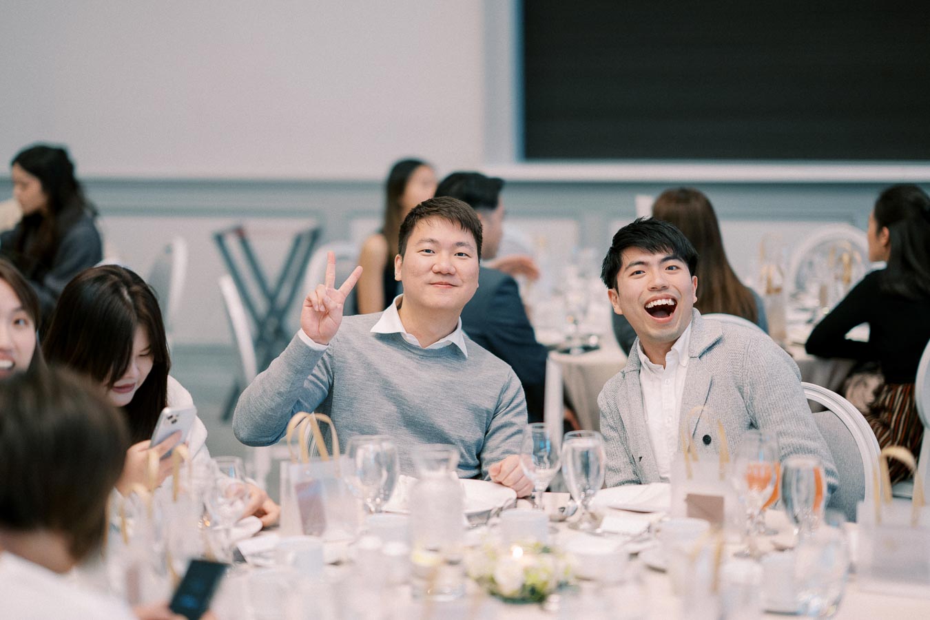 A group of people enjoying a festive meal at a banquet table, with two men in the foreground smiling and making peace signs, surrounded by plates, glasses, and decorative table settings.
