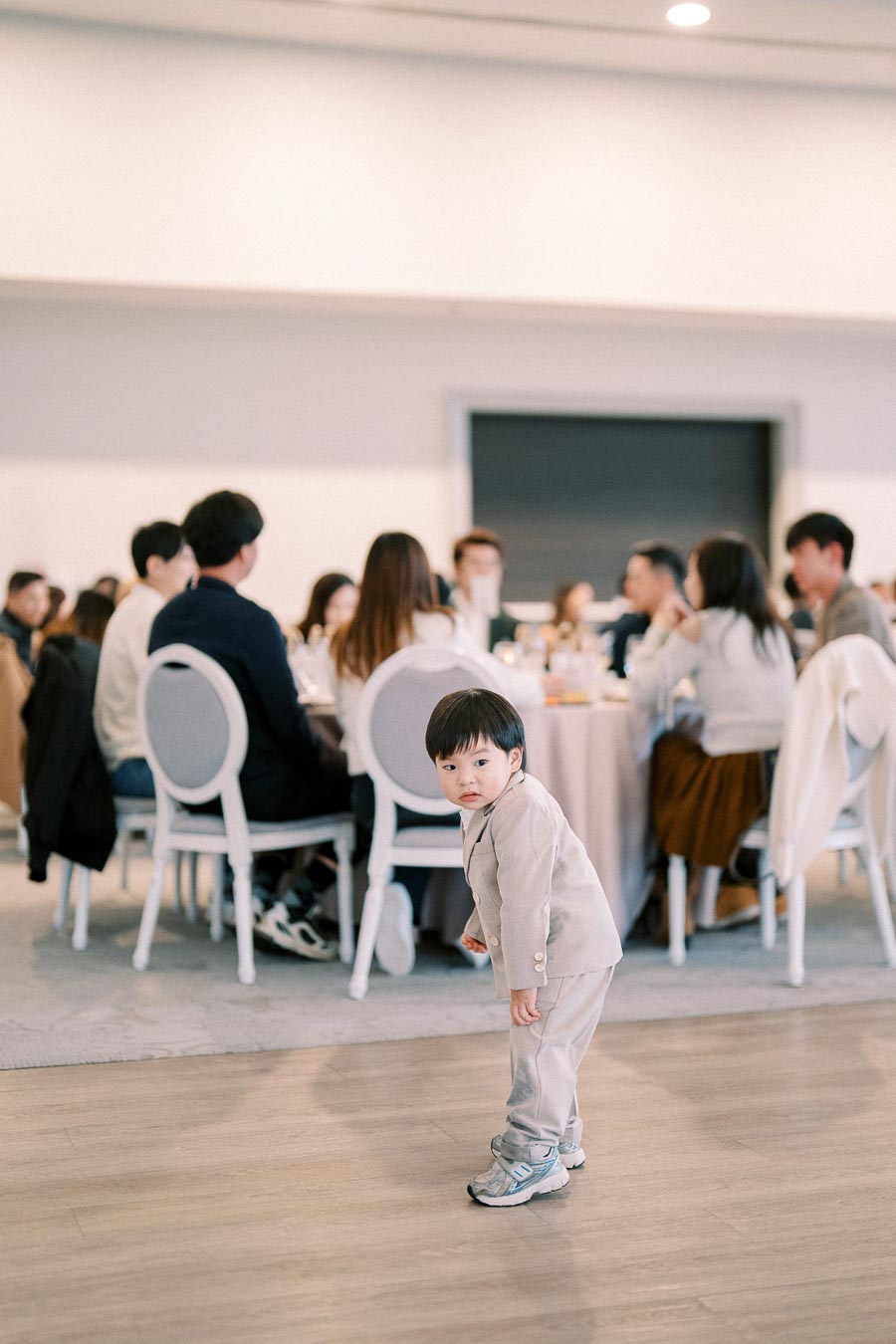 A young child in a light-colored suit stands on a wooden floor at a formal gathering, with people seated at round tables in the background.