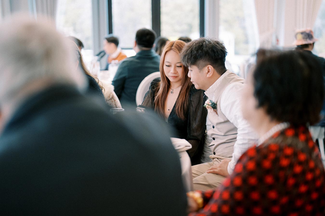 A couple sitting closely together at a formal event, surrounded by other guests, with soft window light illuminating the setting.